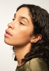 Orecchini a bottone floreali in oro indossati da un modello con capelli ricci, che porta un top verde oliva; texture liscia e piccole dimensioni con dettagli intricati.