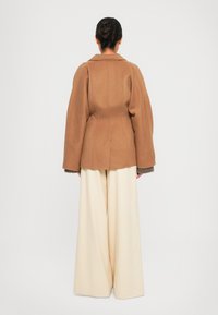 Veste en mélange de laine marron avec un col structuré et des manches larges, associée à un pantalon large en crème fluide. Texture lisse, coupe ajustée.