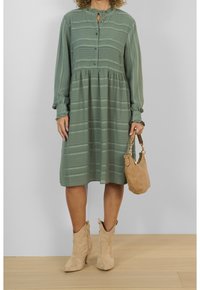 Abito verde, leggero, con maniche lunghe e chiusura a bottoni, caratterizzato da un motivo a righe. Include una vita arricciata e un collo a chiave discreto, abbinato a una borsa beige.