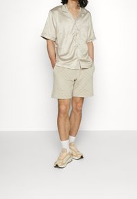 Satinbeige kortärmad skjorta med ficka, matchad med mönstrade shorts. Skorna är ljusa sneakers och vita ankelsockor.