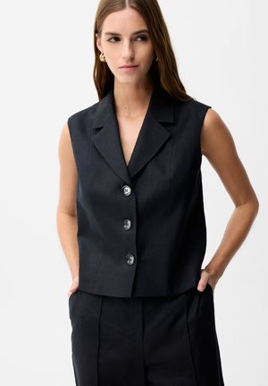 Woman wearing sleeveless black buttoned vest with lapels and matching pants, hands in pockets, against plain light background.