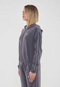 Felpa grigia con zip, maniche lunghe e cappuccio con coulisse. Presenta un cintura elastica aderente e dettagli argento sulle maniche.