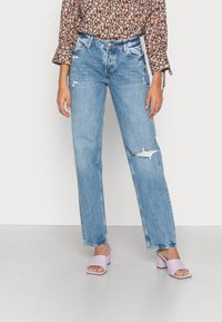 Jeans em azul claro desgastados com corte reto, apresentando rasgos e uma cintura alta, combinados com sandálias de salto bloco rosa.