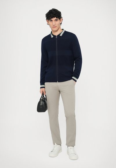 Tmavomodrý sveter na zips s pruhovaným golierom, v kombinácii so svetlosivými nohavicami a bielymi teniskami, drží čiernu kabelku.