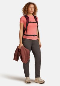 Mochila de forro polar en negro con correas ajustables, llevada sobre una camiseta de manga corta rosa y pantalones grises; sosteniendo una toalla color granate; zapatos de senderismo color beige.