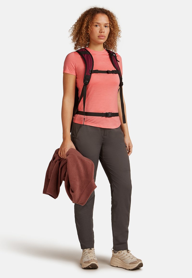 Mochila de forro polar en negro con correas ajustables, llevada sobre una camiseta de manga corta rosa y pantalones grises; sosteniendo una toalla color granate; zapatos de senderismo color beige.