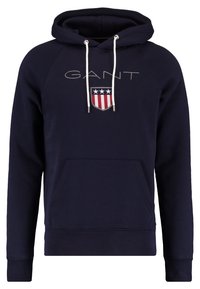 Mörkblå hoodie med vita dragsnören, framficka, "GANT"-text och en sköldlogotyp med tre stjärnor samt röda och vita ränder.