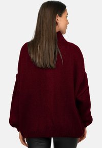 Pull en maille bordeaux à coupe ample, col montant et épaules tombantes. Présente une surface texturée et un ourlet côtelé.