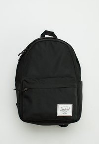 Černý látkový batoh s zaobleným vrškem, přední kapsou na zip a obdélníkovou logovou nášivkou. Má nastavitelné ramenní popruhy a zavěšovací smyčku.