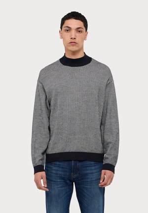 Gestreifter schwarz-weißer Pullover mit hohem Rippkragen, langen Ärmeln und strukturierter Passform. Kombiniert mit blauen Jeans.