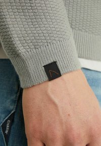 Grå tröja med texturerat stickat mönster, ribbade ärmslut och en svart etikett med en orange logodetalj på ärmen.