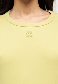 Κοντινό πλάνο ενός ατόμου που φοράει ανοιχτό πράσινο πουκάμισο με στρογγυλή λαιμόκοψη και ανάγλυφο λογότυπο κειμένου "HUGO" στο κέντρο του στήθους.