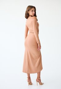 Abito aderente a maniche lunghe in morbido tessuto pesco, con un design drappeggiato in vita e lunghezza a metà polpaccio. Abbinato a décolleté beige.