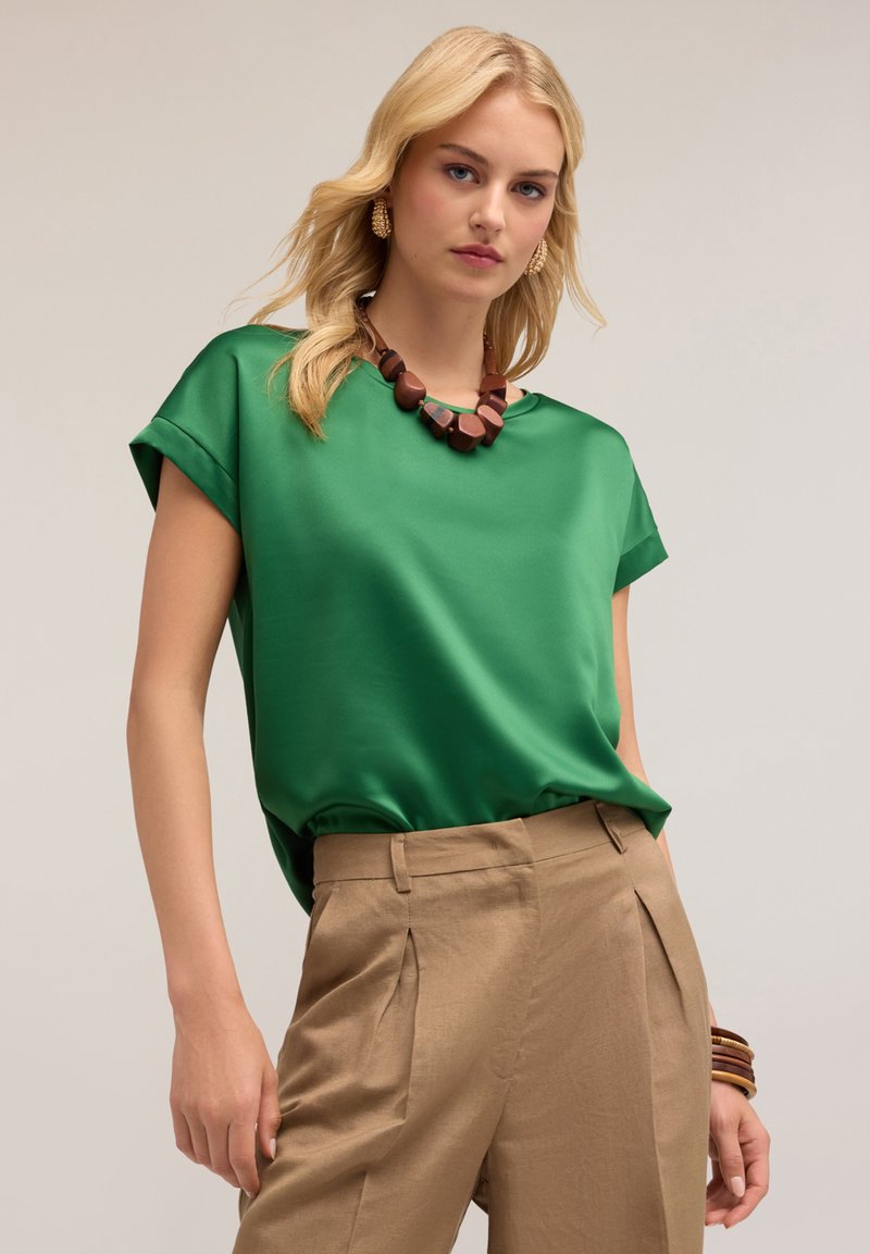 Grön satinklänning med korta ärmar, kombinerad med beige högmidjade byxor. Accessoriserad med ett kraftigt trähalsband och armband.