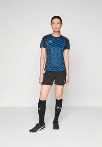 Puma sportstrøye i blått og svart med sikksakkmønster, kombinert med svarte shorts og knelange svarte sokker. Sorte fotballsko fullfører antrekket.