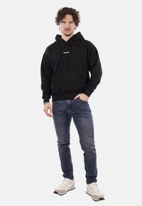Felpa nera con tasca frontale e cappuccio regolabile, abbinata a jeans blu scuro e sneakers beige, mostrata su un modello maschile.