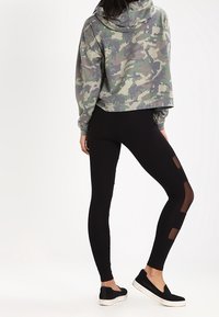 Hoodie court en camouflage dans des tons de vert atténués, associé à des leggings noirs avec des découpes en maille. Des chaussures noires à enfiler complètent le look.