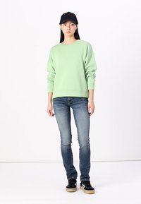 Sweat-shirt surdimensionné vert clair, jeans bleus, baskets noires avec semelles crème, et une casquette noire, présentés sur fond blanc.
