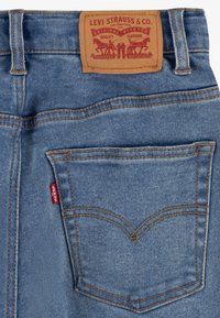 Modré džíny z denimu s hnědou koženou etiketou, která má značkové logo a dvě zadní kapsy s kontrastním prošíváním.