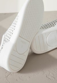 Weiße Sportschuhe mit einer strukturierten Oberschicht und flacher, gemusterter Sohle. Das Markenlabel "NEXT" ist auf den Sohlen sichtbar. Insgesamt glatte Oberfläche.