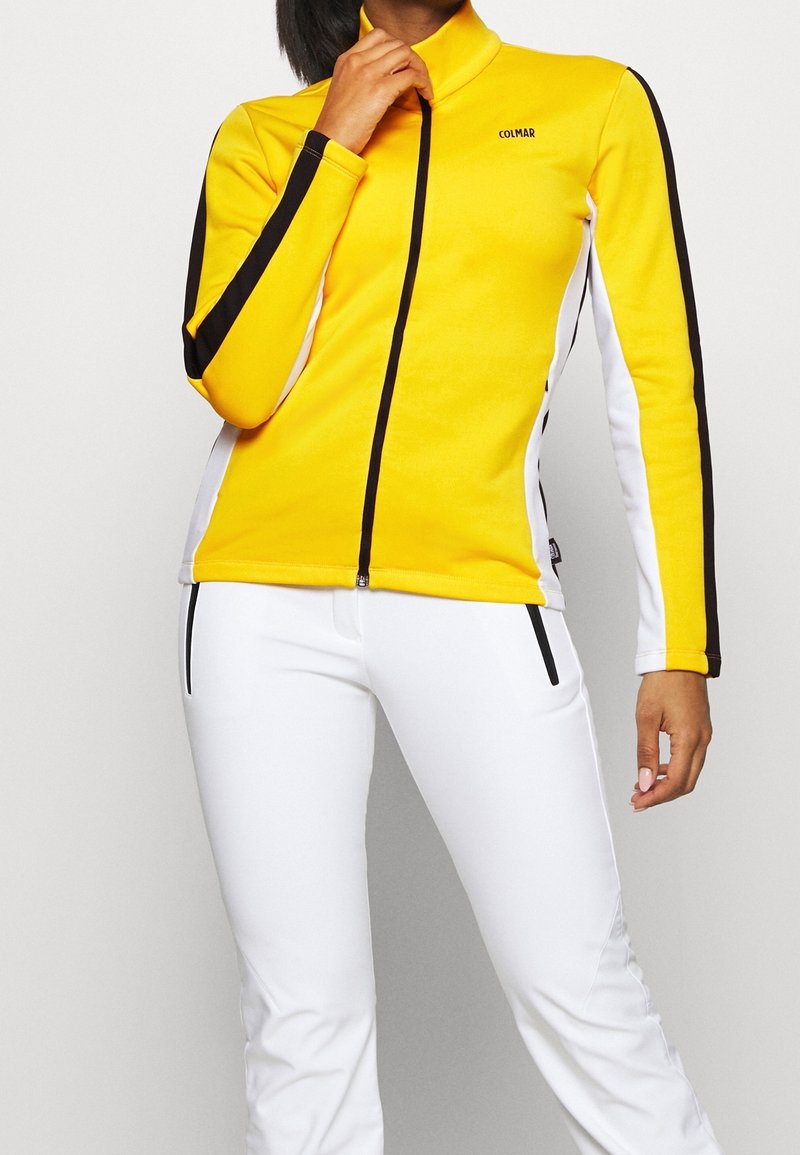 Żółta kurtka zapinana na zamek z czarno-białymi paskami, zestawiona z białymi dopasowanymi spodniami. Kurtka ma wysoki kołnierz i mały detal z logo.