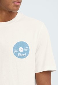 T-shirt met korte mouwen in crèmekleur, met een blauw vinylplaatgrafiek met geluidsgolven en de tekst "Blend" in een casual lettertype.