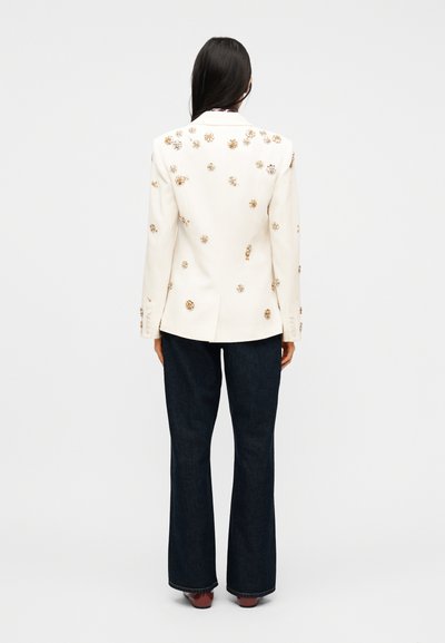 Γυναίκα με μακριά σκουρόχρωμα μαλλιά που φοράει λευκό σακάκι διακοσμημένο με χρυσές λουλουδένιες λεπτομέρειες, σκούρα μπλε παντελόνια φαρδιάς γραμμής και καφέ παπούτσια, γυρισμένη πλάτη.