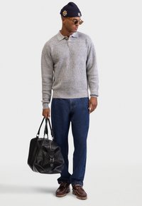 Grå strikket genser, blå jeans, mørkebrune sko og en svart lær duffelbag. Komplettert med en marineblå lue og solbriller.