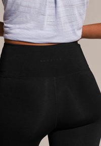 Svarta högmidjade leggings gjorda av de stretcht material, med platta sömmar och en liten logotyp längs midjebandet. Slät textur, minimalistisk design.