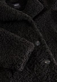 Manteau noir texturé en matière pelucheuse et douce. Doté d'un col et de boutons noirs. Le tissu a un aspect doux et tactile.