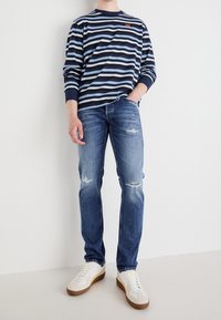 Camisa de rayas azul y marino de manga larga, combinada con jeans claros desgastados y zapatillas blancas con detalles en marrón. Ajuste casual y relajado.