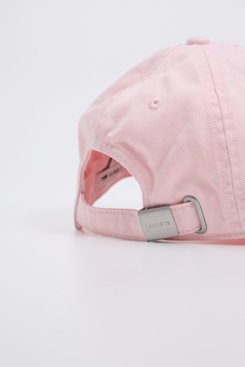 Gorra de algodón rosa con visera curva, correa ajustable y herrajes plateados que muestran la marca "LACOSTE". Textura suave, diseño casual.
