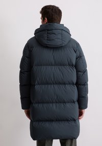 Granatowa pikowana kurtka puchowa z kapturem, posiadająca pikowane sekcje, elastyczne mankiety oraz długość do połowy uda dla dodatkowego ciepła i ochrony.