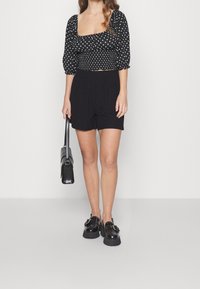 Svart top med prickar och puffärmar, ihop med svarta strukturerade shorts. Modellen håller en svart handväska och har på sig chunky svarta skor.