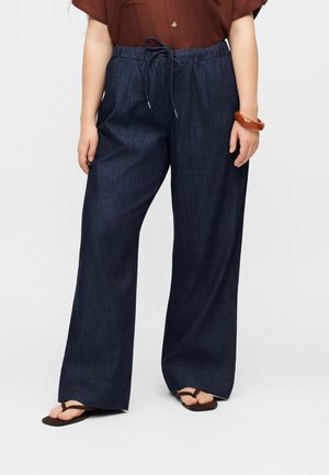 Femme portant un pantalon en jean foncé à jambes larges avec taille à cordon, chemise marron à manches courtes, bracelet marron et tongs noires, debout sur fond blanc.