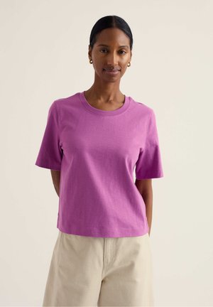 Sötét hajú nő alacsony konttyal, arany karika fülbevalóval, lila rövid ujjú pólóban és bézs nadrágban, egyszínű háttér előtt állva.