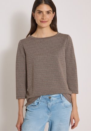 Femme aux longs cheveux bruns portant un haut à manches longues marron et beige à motif pied-de-poule ainsi qu'un jean bleu clair sur un fond uni.