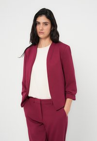 Μπορντό σακάκι με εφαρμοστή γραμμή και μανίκια τριών τετάρτων, σε συνδυασμό με μπλούζα σε κρεμ απόχρωση. Ομαλό ύφασμα, μινιμαλιστικός σχεδιασμός.