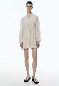 Petites Blush-Kleid mit langen Ärmeln, elastischem Taillengürtel, Bindeschluss am Hals und ausgestellt geschnittenem Saum; getragen mit schwarzen klobigen Flip-Flops.