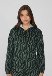 Joven con el cabello largo y castaño que lleva una sudadera de cremallera a rayas de cebra en verde y negro, con bolsillos frontales y un forro de capucha gris.