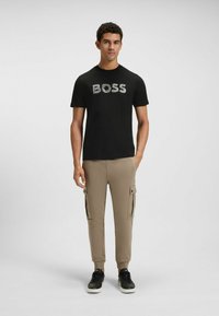 Mustavärinen t-paita, jossa on harmaa "BOSS"-logo, yhdistettynä vaaleanruskeisiin cargo-housuihin, joissa on sivutaskut. Mallilla on päällään mustat lenkkarit.