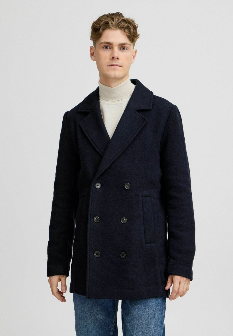 Dobbeltspent marineblå ullkåpe med store svarte knapper, klippelapp og sidelommer, båret over en lys turtleneck og blå jeans.