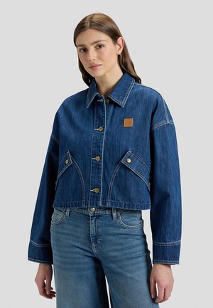 Mujer joven con una chaqueta corta de mezclilla azul oscuro con botones frontales y bolsillos con solapa, combinada con unos vaqueros de tiro alto de color azul claro.