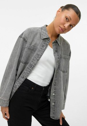 Grijze denim overhemd met een knoopsluiting aan de voorkant, een knoopzak en een relaxte pasvorm. Gecombineerd met een witte tanktop en zwarte broek.