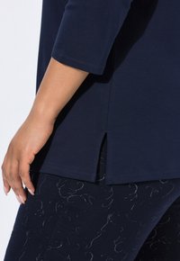Close-up van een hand die rust naast een marineblauw shirt met korte mouwen met een zijsplit en gestructureerde marineblauwe broek met subtiele abstracte patronen.
