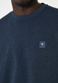 Marineblauwe sweatshirt met geometrische patronen, geribde halslijn en een klein rechthoekig logo-label in marineblauw aan de voorkant.