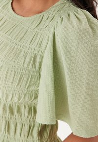 Top verde chiaro realizzato in tessuto strutturato, con un design arricciato, maniche a sbuffo e dettagli di scollatura arricciati.