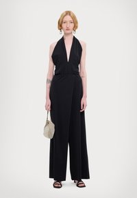 Sort jumpsuit i svart med dyp V-hals, samlet midje, vide ben og glatt tekstur, akkompagnert av en metallisk håndveske og sandaler med stropper.