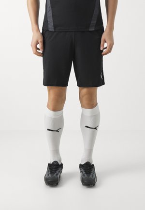 Persoon die zwarte sportshorts draagt, witte kniehoge sokken met zwarte logo's, en zwarte voetbalschoenen, staande tegen een effen lichte achtergrond.