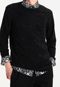 Uomo che indossa un maglione nero a fantasia con un camicia a righe bianca e nera sotto, con colletto e polsini visibili, mani in tasca.